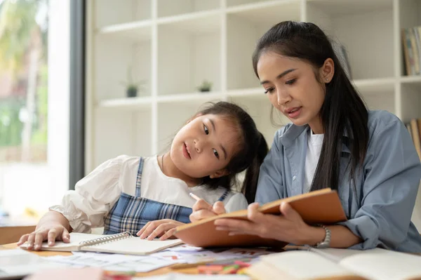 A kind Asian mom is helping with homework and teaching English to her little daughter at home on the weekend. mom and daughter, student and teacher, babysitting