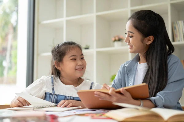 A cheerful Asian female private teacher and a young girl are laughing and enjoying the English class together. Kids education concept