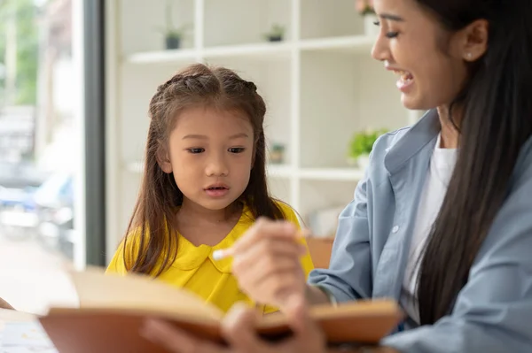 A pretty Asian preschool girl is studying and reading texts in a book with a kind and caring female teacher. Learning, studying, child development, parenting