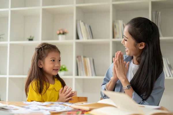 A caring and kind Asian female teacher is clapping her hands with a little girl, feeling proud of her student, and enjoying the class together.