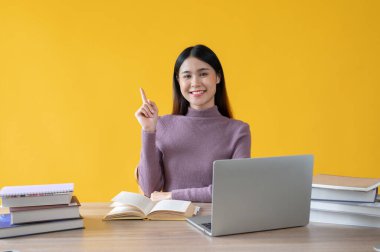 Güzel ve zeki bir Asyalı genç kız üniversite öğrencisi çalışma masasında oturuyor, parmağını kaldırıp gülümsüyor ve kameraya bakıyor. izole edilmiş sarı arkaplan.