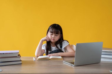 Çalışma masasında izole edilmiş sarı bir arka planda kitap okumaya odaklanan sevimli, genç bir Asyalı kız. Anaokulu, ilkokul öğrencisi, boş zaman ve hobi, çocuklar.