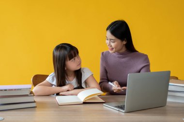 Sevimli genç bir Asyalı kız, bir bayan öğretmenle ders çalışıyor, aynı masada oturuyor. İzole edilmiş sarı arka plan. özel öğretmen, ev okulu, eğitim konsepti