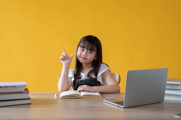 一位可爱的年轻亚洲女孩坐在自己的书桌前 桌上放着课本和笔记本电脑 指尖向上 微笑着看着相机 孤立的黄色背景 — 图库照片