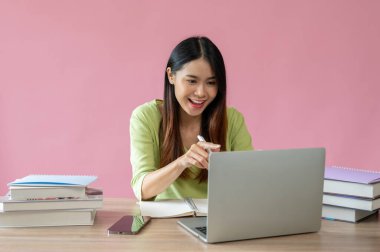 Çekici ve neşeli bir Asyalı kız üniversite öğrencisi çalışma masasında dizüstü bilgisayarında ders alıyor. İzole edilmiş pembe arka plan. e-öğrenme, çevrimiçi çalışma