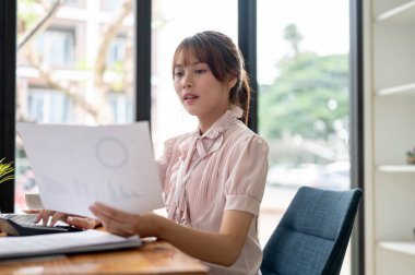Profesyonel bir Asyalı iş kadını ya da kadın finans asistanı finansal bir raporu inceliyor, satış verilerini analiz ediyor ve ofisindeki masasında evrak işleriyle uğraşıyor..