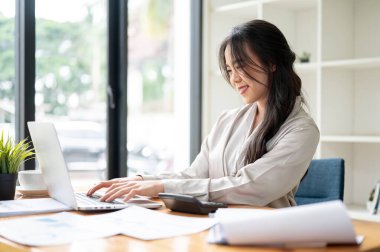 Çekici ve profesyonel bir Asyalı iş kadını, ofisteki masasında çalışan bir dizüstü bilgisayarla işine odaklanıyor..