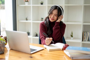 Güzel ve rahat, kırmızı kazaklı Asyalı bir kadın, kahve dükkanında uzaktan çalışırken kulaklıklarıyla müzik dinliyor..
