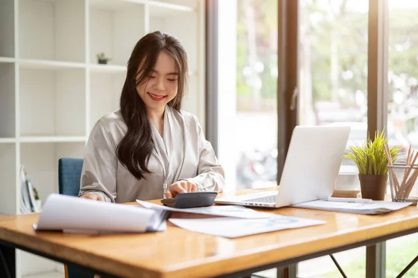 Güzel ve profesyonel bir Asyalı iş kadını, satış verilerini hesaplamak ve ofisindeki masasındaki işine odaklanmak için hesap makinesi kullanıyor..