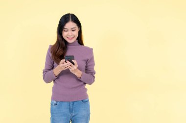 Güzel ve mutlu bir Asyalı kadın, izole edilmiş sarı arka planda dururken akıllı telefonunu kullanıyor. metin, sohbet, mesaj, uygulama, sosyal medya