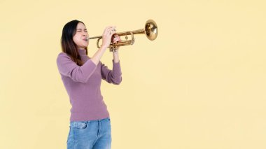 Güzel ve yetenekli bir Asyalı kadın izole edilmiş sarı bir stüdyo arkasında dururken trompet çalıyor. müzisyen, müzik, enstrüman