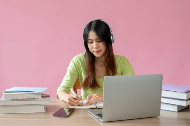 Genç ve güzel bir Asyalı kız üniversite öğrencisi çevrimiçi bir derse çalışmaya ya da çalışma masasında soyutlanmış pembe bir arka plana karşı ödev yapmaya odaklanır..