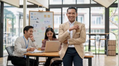 Yakışıklı ve başarılı bir Asyalı erkek CEO ya da iş adamı elinde kahve fincanıyla kameraya gülümserken, çalışanları toplantı odasında bir masada çalışıyor..
