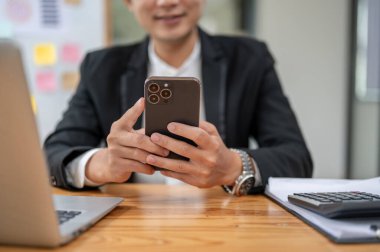 Ofisteki masasında akıllı telefonunu kullanan mutlu ve profesyonel bir Asyalı iş adamının yakın plan görüntüsü. metin, sohbet, SMS, e-posta, uygulamalar