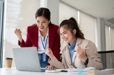 İki neşeli ve heyecanlı Asyalı iş kadını mutlu yüzlü bir dizüstü bilgisayar ekranına bakıyor ve projelerinin başarısını ofiste birlikte kutluyorlar. takım çalışması konsepti