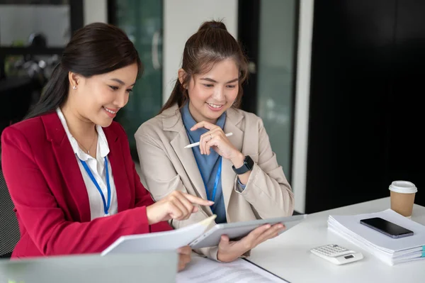 Two beautiful Asian businesswomen are examining a project\'s details and working on it together at a table in the office. co-working, teamwork, coworker