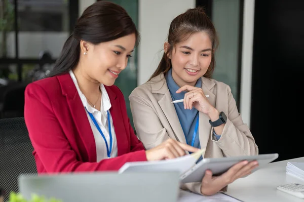 Two professional and attractive Asian businesswomen are examining a project\'s details and working on it together at a table in the office. co-working, teamwork, coworker