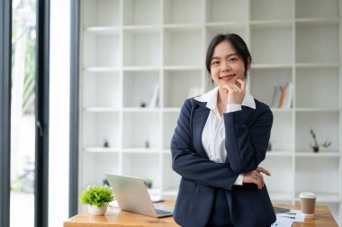 Kendine güvenen ve çekici Asyalı bir iş kadını ya da resmi takım elbiseli bir kadın avukat kameraya gülümsüyor ve modern ofisinde duruyor..