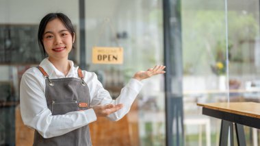 Güzel ve gülümseyen Asyalı bir bayan kafe çalışanı ya da önlüklü küçük bir işletme sahibi kafesinin girişinde dostça bir jest yapıyor..
