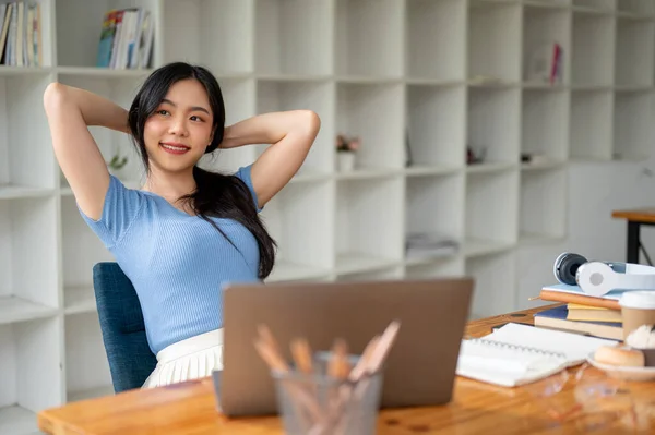 A beautiful and happy Asian woman relaxes, puts her hands behind her head, and looks out the window with a happy face at her study table, enjoying a carefree moment at home after work.