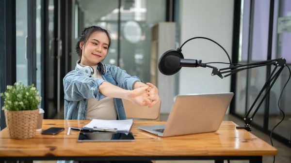 A beautiful and happy Asian female radio host or content creator relaxes and stretches her arms at her desk after finishing her live radio show in the studio.