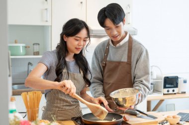 Genç ve güzel bir Asyalı kız arkadaşı mutfakta erkek arkadaşıyla krep yapıyor, spatulayla krepleri çeviriyor ve hafta sonunun keyfini çıkarıyor..