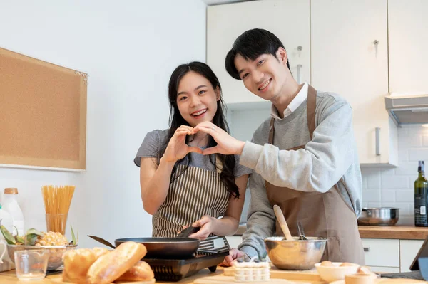 A loving young Asian couple is making a heart hand sign together while making pancakes in the kitchen. Relationship, cooking dates, romantic life, home cooking
