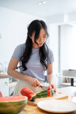 Genç ve güzel bir Asyalı kız doğrama tahtasıyla karpuz kesmeye ve mutfakta sağlıklı atıştırmalıklarını hazırlamaya odaklanıyor..