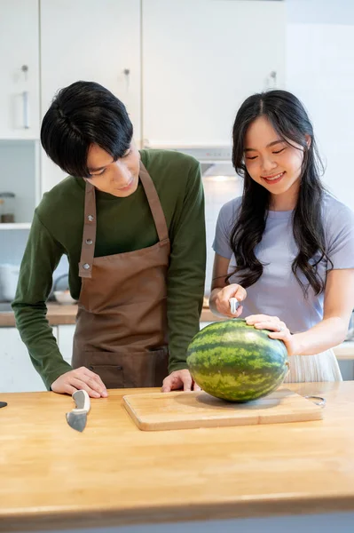 A lovely young Asian couple is preparing healthy food in the kitchen together, cutting a watermelon on a chopping board, and spending happy, quality time together on the weekend.