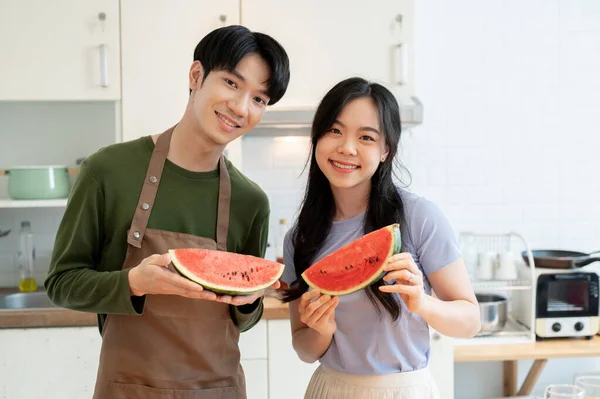 A lovely and happy young Asian couple is holding a slice of watermelon and enjoying cooking healthy meals in the kitchen together.