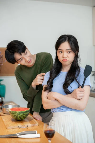 A cute, clingy young Asian boyfriend is asking for forgiveness from his angry girlfriend after arguing while cooking in the kitchen together.