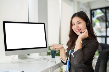 Güzel bir Asyalı kadın çağrı merkezi operatörü ya da kulaklıklı müşteri destek servisi temsilcisi ofisindeki masasında oturuyor. Bilgisayar beyaz ekran modeli. Teknik destek, satış temsilcisi, yardım hattı.