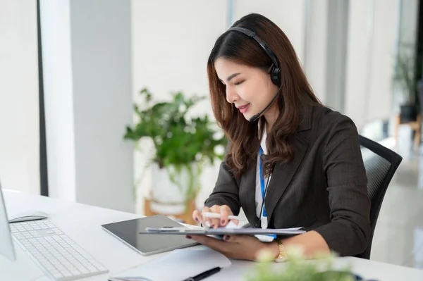 stock image A beautiful Asian female call centre operator or customer support service agent with a headset is working at her desk in the office.