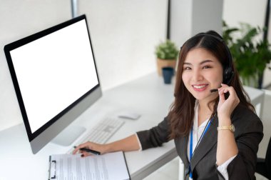 Çekici bir Asyalı kadın çağrı merkezi operatörü ya da kulaklıklı müşteri destek servisi temsilcisi ofisindeki masasında oturuyor. Bilgisayar beyaz ekran modeli. tele pazarlama, teknik destek