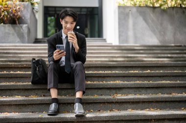 Mutlu ve yakışıklı bir Asyalı genç işadamı ya da erkek ofis çalışanı binanın önündeki merdivenlerde otururken akıllı telefonunu kullanıyor ve kahvesini yudumluyor. Yemek molası, şehir yaşam tarzı.