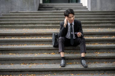 Stresli ve yorgun Asyalı bir işadamı ya da erkek ofis çalışanı gözlüklerini çıkarıp binanın önündeki merdivenlere oturur..