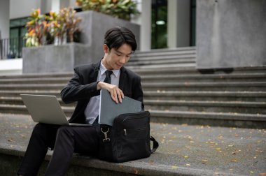 Zeki ve yakışıklı Asyalı genç bir işadamı ya da erkek ofis çalışanı şehrin önündeki merdivenlerde otururken uzaktan çalışıyor..