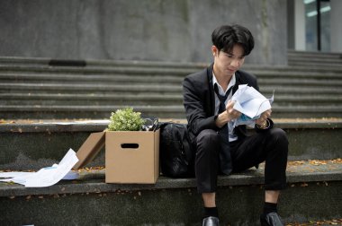 Sinirli ve öfkeli genç Asyalı işsiz bir işadamı işini kaybettikten sonra şirket ofisinin önünde bir kutu kişisel eşyası ile merdivenlerde otururken kağıtları buruşturuyor..