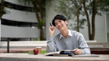 Yakışıklı ve mutlu bir Asyalı erkek üniversite öğrencisi kitap okumaktan ve kampüs parkında sınava hazırlanmaktan zevk alıyor..