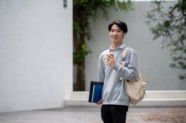 Sırt çantasıyla kampüs binasında elinde kahve fincanıyla gezen yakışıklı ve genç Asyalı bir erkek öğrenci. Üniversite, eğitim, üniversite