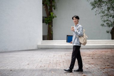 Sırt çantasıyla kampüs binasında elinde kahve fincanıyla gezen yakışıklı, genç Asyalı bir erkek öğrenci..