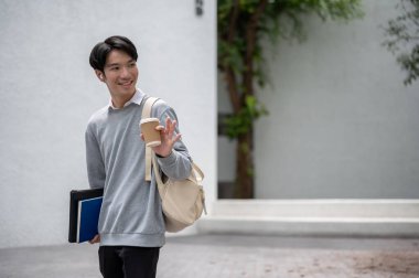 Yakışıklı ve gülümseyen bir Asyalı erkek öğrenci. Sırt çantası elinde kahve fincanıyla kampüs binasında yürüyor..