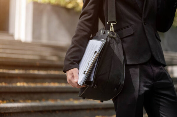 Cropped image of a professional businessman or lawyer in a formal black suit with his briefcase and clipboard walking down the stairs in the city.