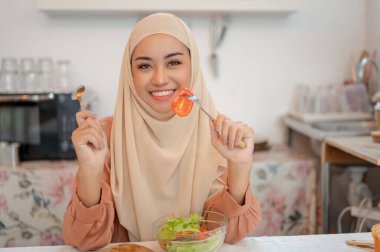 Güzel, Asyalı, tesettürlü Müslüman bir kadın mutfaktaki bir yemek masasında sağlıklı salata kasesini yemekten zevk alır. Sağlıklı yemek, diyet, refah yaşam tarzı