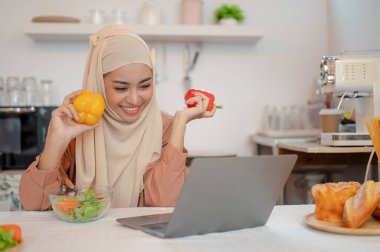 Mutlu ve güzel bir Asyalı Müslüman kadın mutfaktaki yemek masasında dizüstü bilgisayarı üzerinde çalışıyor. Evde çalışmak, Gıda blogcusu, ev hayatı