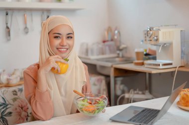 Güzel bir Asyalı Müslüman kadın mutfaktaki yemek masasında laptopuyla çalışırken sağlıklı salatasını yiyor. Evden çalışma, serbest çalışma, uzaktan çalışma, ev hayatı, yemek blogcusu