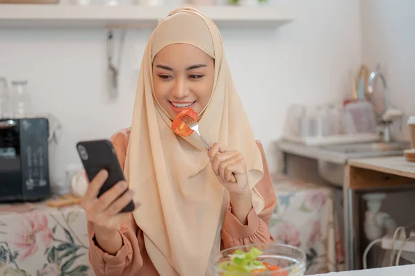 ヒジャブを着ている美しいアジアのイスラム教徒の女性は 彼女のキッチンのダイニングテーブルで彼女のスマートフォンを使用しながら 彼女の健康的なおいしいサラダボウルを食べることを楽しんでいます — ストック写真