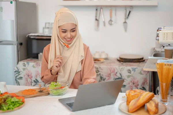 ヒジャーブを着ている美しいアジアのイスラム教徒の女性は 彼女の台所のダイニングテーブルで彼女のラップトップを使用しながら 彼女の健康的なおいしいサラダボウルを食べています 家庭生活や家庭生活から — ストック写真