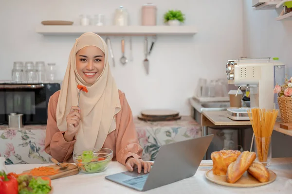 美しいアジアのイスラム教徒の女性がキッチンの食卓でノートパソコンで働きながら健康的なサラダを食べています フリーランス リモートワーキング ライフスタイル — ストック写真