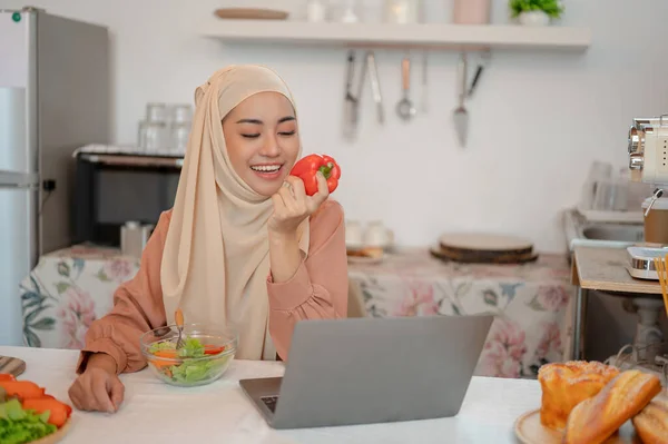ハッピーで美しいアジアのイスラム教徒の女性は キッチンのダイニングテーブルで彼女のラップトップで働いている間 彼女の健康的なサラダを食べることを楽しんでいます 自宅から仕事 食品ブロガー ライフスタイル — ストック写真